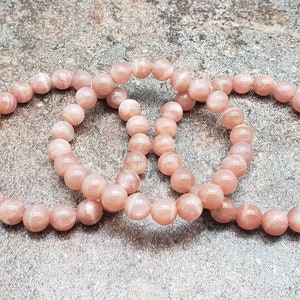 May include: Three pink sunstone beaded bracelets, each with a round shape, are arranged in a circle on a gray textured surface.