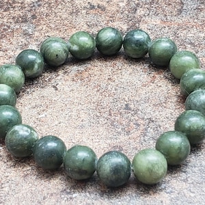 May include: A green jade bead bracelet. The bracelet features round, polished beads in varying shades of green, creating a natural, earthy aesthetic. The beads are strung together, forming a circular shape. The bracelet is displayed on a textured, neutral-toned surface.