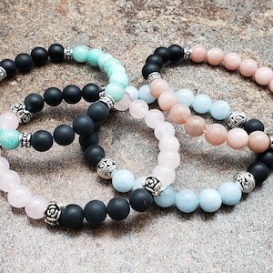 May include: Four bracelets made of natural stones. The bracelets are layered on top of each other. The bracelets are made of black, light blue, pink, and green stones. Each bracelet has silver accents.