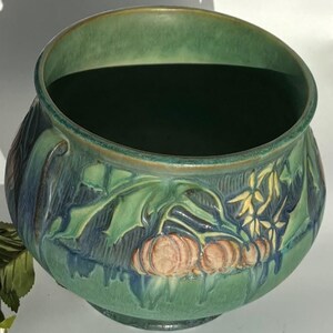May include: A green ceramic planter with a raised design of leaves and pumpkins. The planter has a wide, round opening and a slightly flared base.