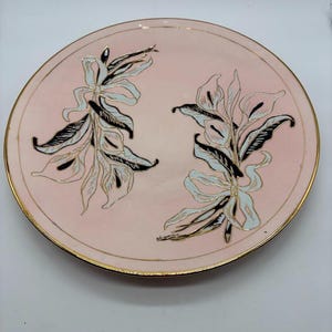 May include: A round, pink plate with a gold rim. The plate features two floral designs in black, white, and gold. The floral designs are symmetrical and placed on either side of the plate's center.