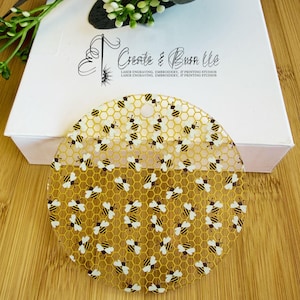 May include: A circular clear acrylic ornament featuring a repeating pattern of yellow and black bees over a gold honeycomb grid. The ornament has a small hole at the top for hanging and is positioned in front of a white box with the text Create & Burn LLC.