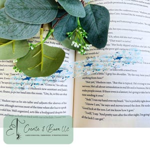 May include: A decorative washi tape roll with a light blue background and a pattern of small blue whales. The tape is placed on an open book with green leaves and white flowers.