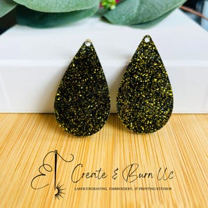 May include: Two teardrop-shaped earrings with a black and gold glitter design. Each earring has a small hole at the top for hanging. The earrings are displayed on a wooden surface. The text "Create & Burn Uc" is visible.