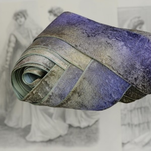 Può includere: Nastro di velluto arrotolato nelle tonalità del blu, grigio e verde oliva. Il nastro ha un aspetto strutturato ed è strettamente arrotolato, rivelando un lato inferiore azzurro. Sullo sfondo c'è un'illustrazione vintage.