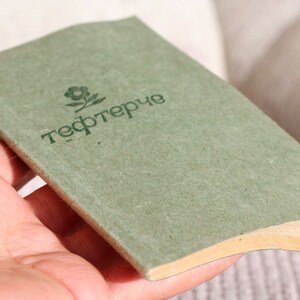 May include: A green notebook with the word "тефтерче" printed on the cover. The notebook has a brown spine and is held in a hand.