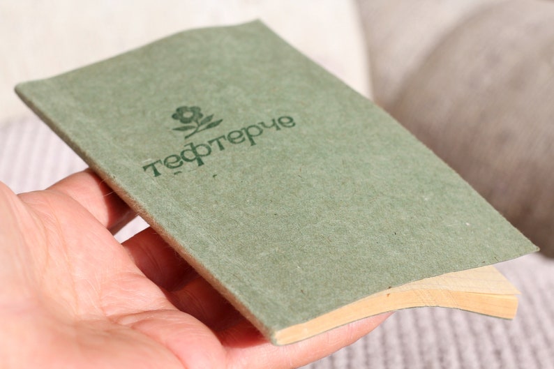 May include: A green notebook with the word "тефтерче" printed on the cover. The notebook has a brown spine and is held in a hand.
