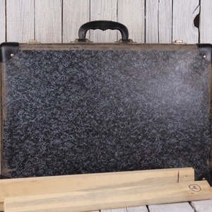 May include: A vintage black suitcase with a speckled pattern and a black handle. The suitcase is sitting on a wooden surface.