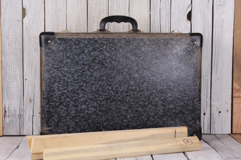 May include: A vintage black suitcase with a speckled pattern and a black handle. The suitcase is sitting on a wooden surface.