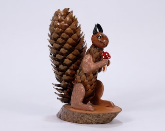 May include: A brown squirrel figurine made from pine cones, holding a red and white mushroom. The squirrel is standing on a wooden base.