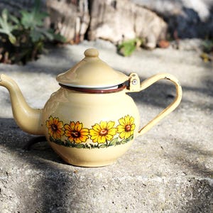 Könnte beinhalten: Eine gelbe Emailleteekanne mit braunem Rand und einem Blumendesign aus Sonnenblumen. Die Teekanne hat einen Deckel und einen Griff.