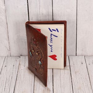May include: A brown leather journal with an intricate design on the cover. The journal is open to a blank page with the words "I Love You" written in blue ink.