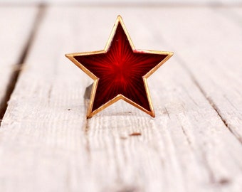 May include: A five-pointed star ring featuring a deep red translucent enamel inlay with a sunburst pattern, encased in a thin gold-toned metal frame. The star is mounted on a simple metal band that extends from the back of the star shape.
