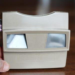 May include: A vintage tan-colored View-Master viewer. The viewer has two eye pieces and a slot for inserting the View-Master reels. The viewer is held by a hand. The viewer is made of plastic and has a rectangular shape.