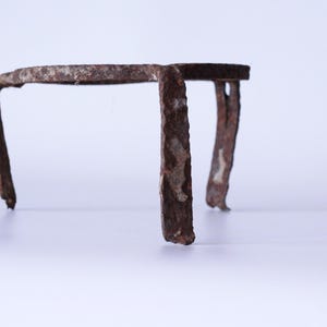 Può includere: Un piccolo supporto in metallo arrugginito con tre gambe e una parte superiore curva. Il supporto è marrone con segni visibili di corrosione, che suggeriscono l'età e l'esposizione agli agenti atmosferici. Il design è semplice e funzionale.