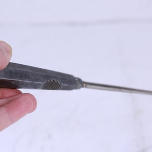 Puede incluir: Una herramienta de metal plateado con un mango largo y un bucle cuadrado en el extremo. El mango tiene un agarre texturizado.