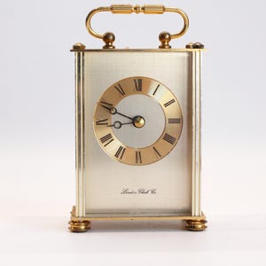 Peut inclure: Une horloge de voyage rectangulaire dorée avec une poignée. Le cadran de l'horloge comporte des chiffres romains et des aiguilles noires. Les mots "London Clock Co." sont imprimés sur le cadran. L'horloge a des pieds dorés.