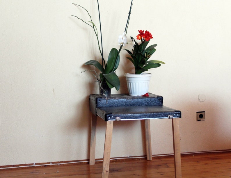 May include: A black wooden table with two shelves, holding two potted plants. The top shelf has a white orchid plant with white flowers. The bottom shelf has a red flowering plant in a white pot.