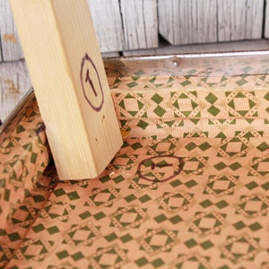 May include: Close-up of the inside of a vintage suitcase with a brown and green geometric pattern. The suitcase is lined with a brown and green patterned paper. The numbers "1" and "5" are circled in black marker.