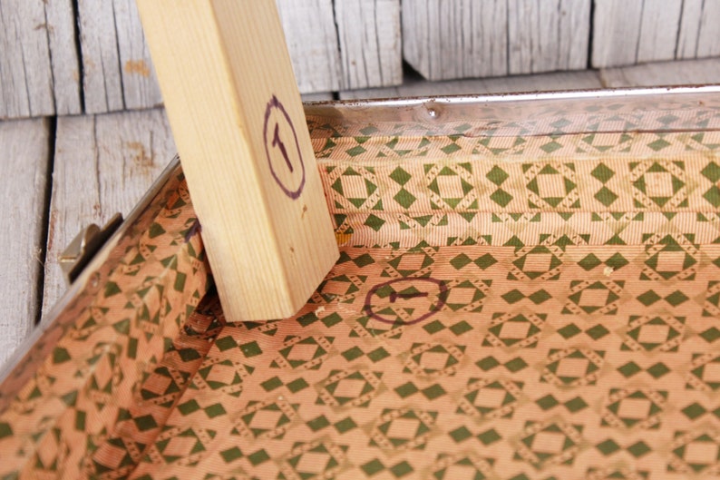 May include: Close-up of the inside of a vintage suitcase with a brown and green geometric pattern. The suitcase is lined with a brown and green patterned paper. The numbers "1" and "5" are circled in black marker.