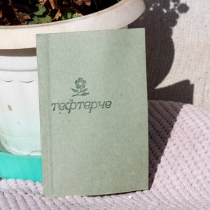 May include: A green notebook with a small flower design and the word "тефтерче" printed on the cover.