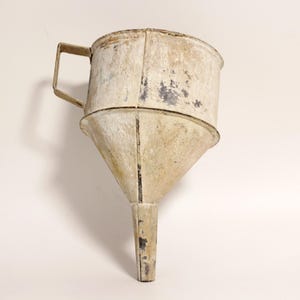 Può includere: Un imbuto antico in metallo con manico. L'imbuto è realizzato in metallo di colore chiaro con segni di usura visibili, tra cui scolorimento e ruggine. Il design include una parte superiore ampia, un corpo conico e un beccuccio stretto.
