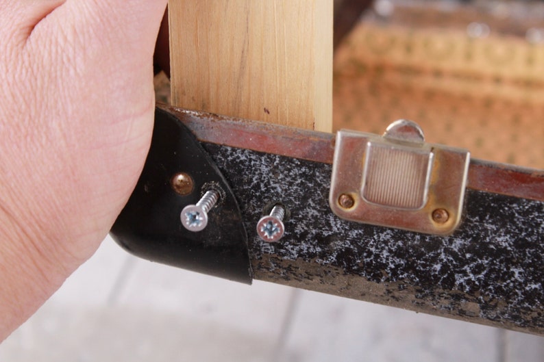 May include: Close-up of a black suitcase with a metal latch and two screws. The suitcase is being held by a hand.