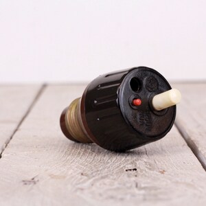 May include: A vintage electrical plug. It has a dark brown body, a cream-coloured cylindrical component, and a small red button. The plug is on a light-coloured wooden surface, suggesting a retro or antique aesthetic.