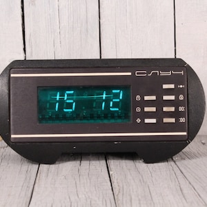 May include: A black digital clock showing the time 15:12 in green. The clock has a rectangular display and buttons on the right. The word "СЛУЧ" is printed on the top. The clock is likely from the Soviet era.
