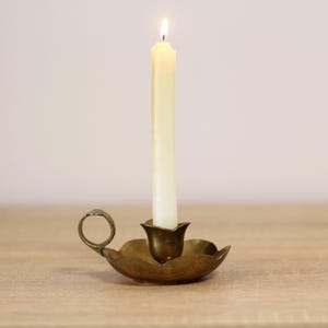 Puede incluir: Un candelero de latón con una base en forma de hoja y un asa. Una vela blanca está encendida en el candelero.