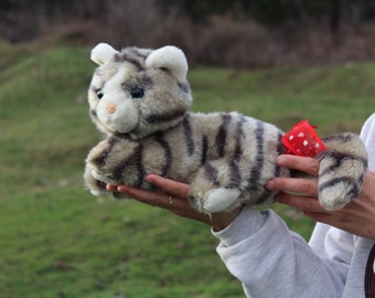 May include: A person holding a grey and white striped plush cat toy with a red bow. The toy has a white face with black eyes and a pink nose.