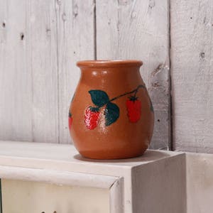 Könnte beinhalten: Eine braune Keramikvase mit einem bemalten Design aus grünen Blättern und roten Erdbeeren.