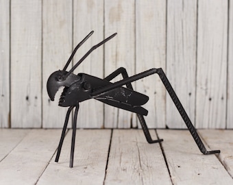 May include: A black metal sculpture of a grasshopper made from recycled metal parts. The sculpture is posed in a jumping position with its legs extended.
