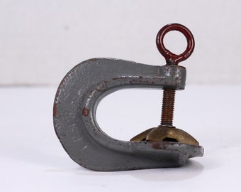 May include: A gray metal C-clamp with a brass screw and a red ring on the top. The clamp is used for holding objects together.