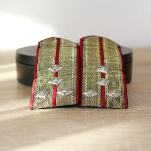 Puede incluir: Par de charreteras militares vintage. Las charreteras presentan un fondo dorado con rayas rojas y adornos plateados. Se muestran sobre una superficie de madera.