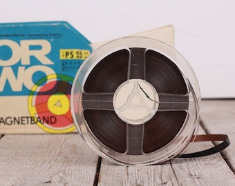 Könnte beinhalten: Eine durchsichtige Kunststoffspule mit braunem Audioband und einem schwarzen Bandanfang. Die Spule ist mit "PS 25" beschriftet und liegt auf einer Holzoberfläche. Die Spule befindet sich neben einer blau-weißen Schachtel mit der Aufschrift "ORWO MAGNETBAND".