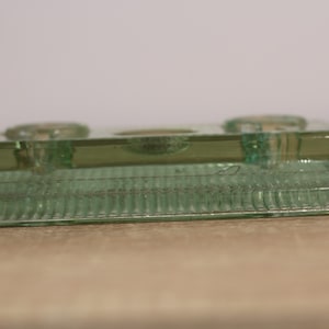 Puede incluir: Un portavelas rectangular de vidrio verde claro con tres hendiduras circulares. La base tiene un diseño texturizado y acanalado. El portavelas está sobre una superficie marrón claro.