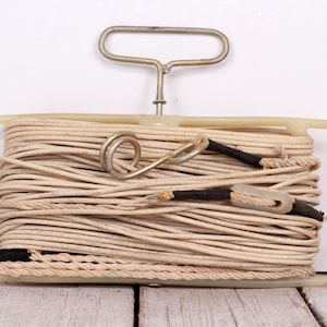 Peut inclure: Une bobine de corde beige avec un cordon noir enroulé autour. La corde est maintenue dans un support en plastique avec une poignée en métal.