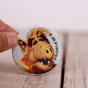 May include: A round button with a cartoon image of ALF, the alien from the TV show. The button is white with a blue background and the text "NO PROBLEMS" written around the edge.