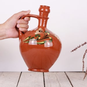 May include: A hand holds a brown ceramic pitcher with a floral design. The pitcher has a wide base and a narrow neck with a spout. The floral design is painted in white, green, and yellow.