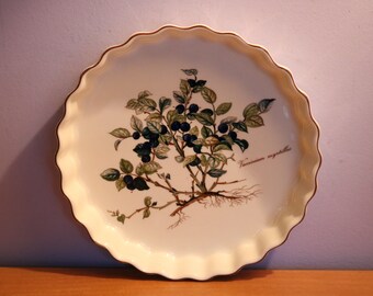 May include: A white ceramic baking dish with a fluted rim and a botanical illustration of blueberries and leaves. The dish has a gold trim and the text "Vaccinium myrtillus" is printed on the bottom right.