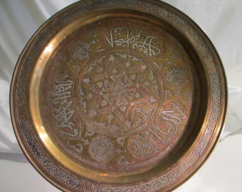 May include: A large, round, brass tray with intricate engraved designs. The center features a six-pointed star surrounded by Arabic calligraphy. The tray has a raised rim with a decorative border.