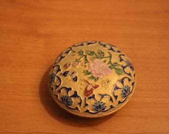 May include: A round, gold-colored trinket box with a blue, green, and pink floral design. The box is decorated with butterflies and flowers.