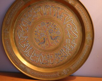 May include: A round, gold-colored metal tray with intricate engraved designs. The tray features Arabic calligraphy and a central motif of a tree with birds.