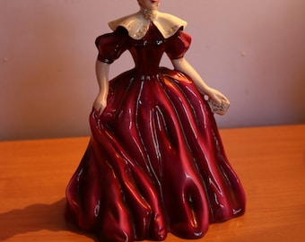 May include: A porcelain figurine of a woman wearing a burgundy dress with a white collar and a gold crown. The dress has a flowing skirt and the woman is holding a small object in her hand.