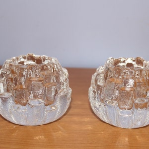 May include: Two clear glass candle holders with a textured, geometric design. The holders are round and have multiple openings for candles.