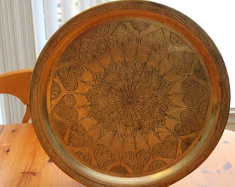 May include: A round, brass serving tray with an intricate, engraved design of hearts and geometric patterns. The tray has a raised rim and a polished finish.