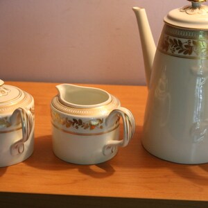 May include: A white porcelain tea set with gold trim. The set includes a teapot, creamer, and sugar bowl. The teapot has a tall, slender body and a spout on the side. The creamer and sugar bowl have rounded bodies and handles.