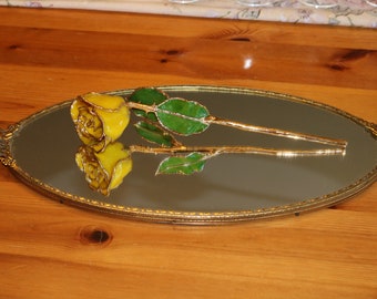 May include: A single yellow rose with green leaves is displayed on a gold oval mirror tray.