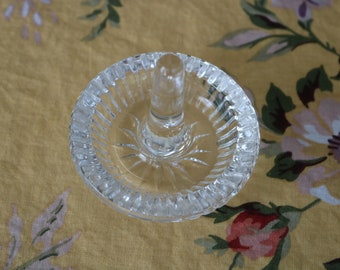 May include: A clear glass ring holder featuring a circular base with a ribbed, scalloped edge and a central vertical post. The glass surface is transparent and smooth, reflecting light across its faceted design, providing a functional display.